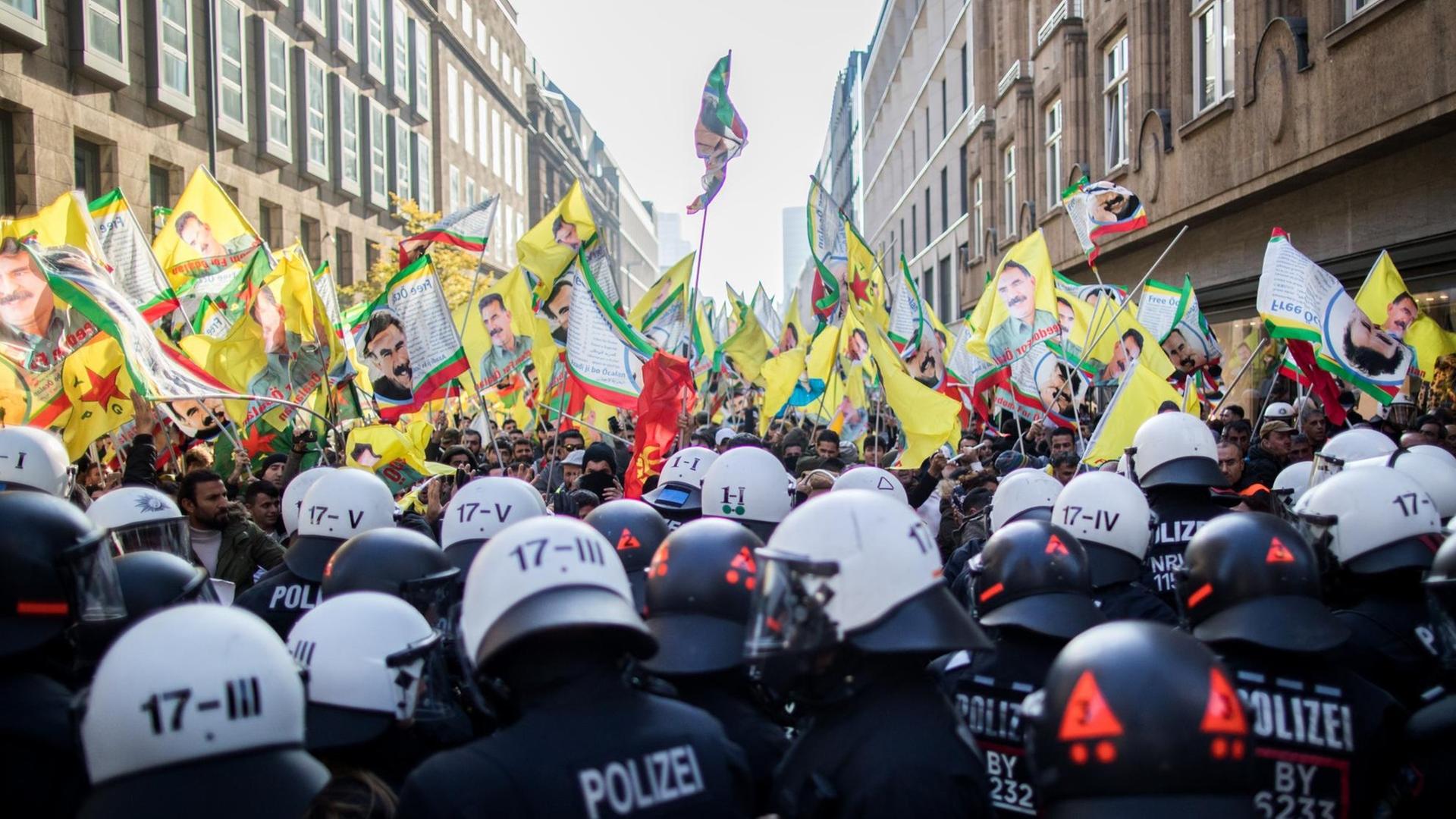 Im Vordergrund viele Polizisten mit weißen Helmen. Dahinter die Demonstranten mit vielen gelben Fahnen, auf denen Öcalan zu sehen ist.