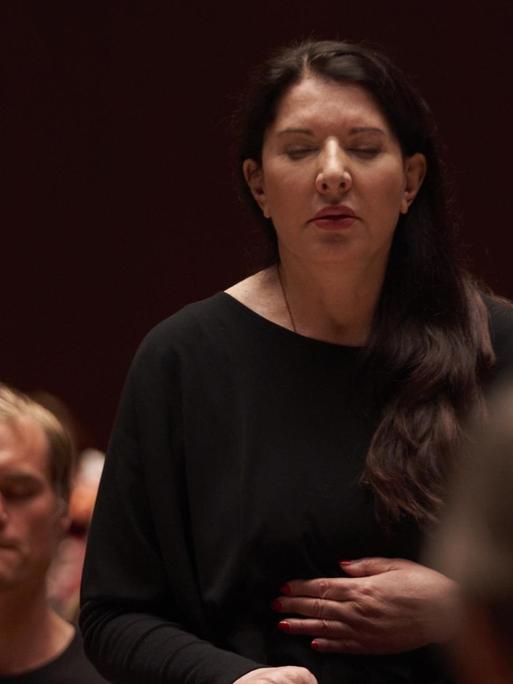 Marina Abramovic steht im Publikum, Hand auf dem Bauch und Augen geschlossen. Sie trägt schwarz.