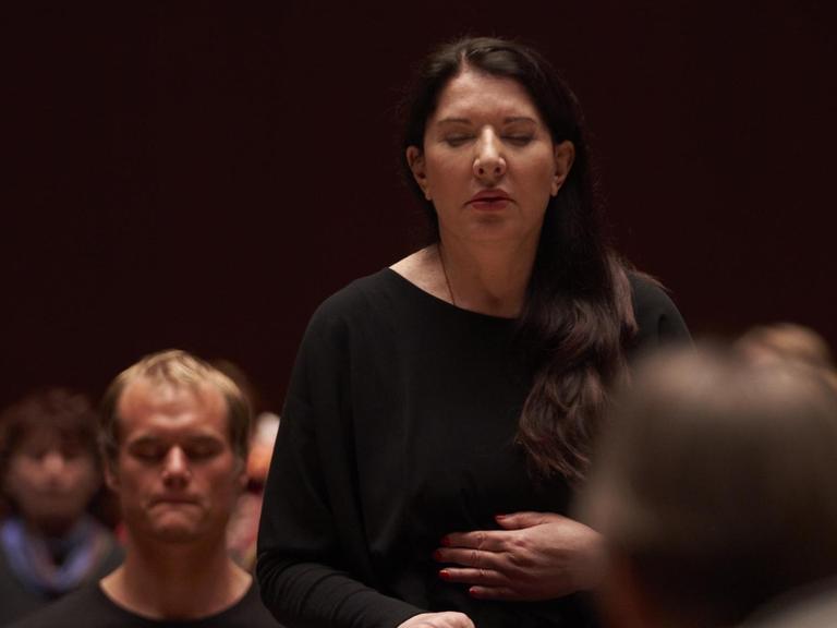 Marina Abramovic steht im Publikum, Hand auf dem Bauch und Augen geschlossen. Sie trägt schwarz.