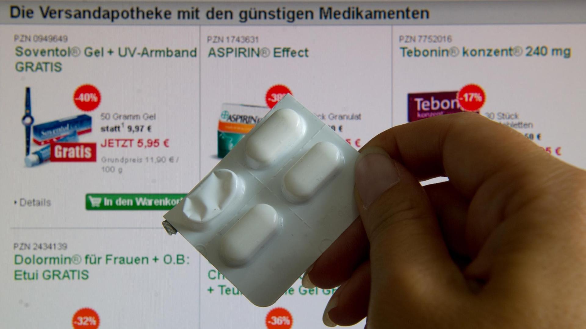 Eine Hand hält einen Blister mit Tabletten vor einen Bildschirm, auf dem ein Teil des Internetauftritts einer Online-Apotheke zu sehen ist.