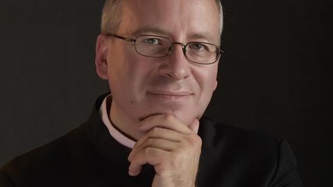 Ein Mann mit Brille schaut in die Kamera, wobei er eine Hand an sein Kinn gelegt hat.
