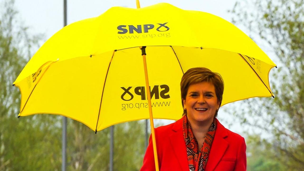 Das Foto zeigt die schottische Regierungschefin Nicola Sturgeon. Sie trägt einen roten Mantel und steht unter einem gelben Regenschirm mit dem Parteilogo der Schottischen Nationalpartei.