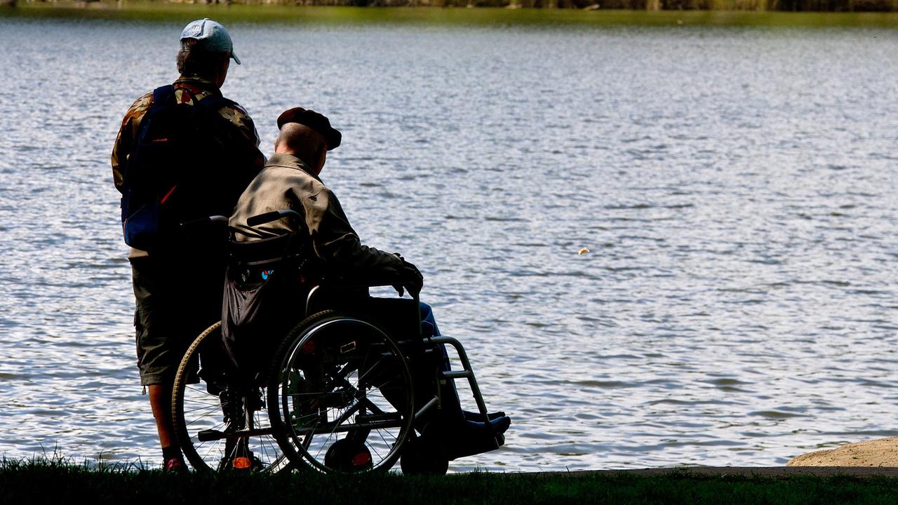 Ein Mann begleitet einen älteren Herren in einem Rollstuhl um einen See.