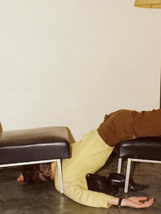 Ein Mann in brauner Hose und gelbem Hemd, liegt halb unter und über einem Sesselstuhl.