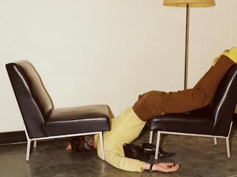 Ein Mann in brauner Hose und gelbem Hemd, liegt halb unter und über einem Sesselstuhl.