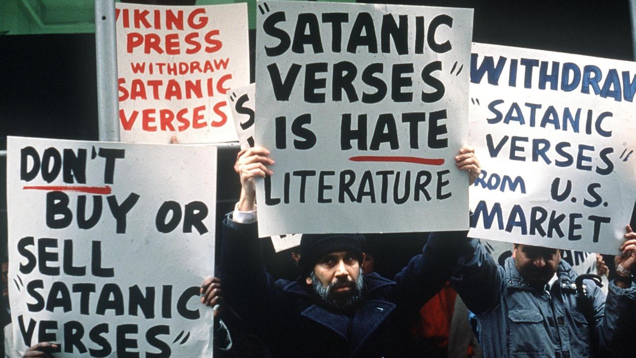 Mehrere muslimische Demonstranten halten Plakate hoch, auf denen das Buch "Satanische Verse" verurteilt wird.