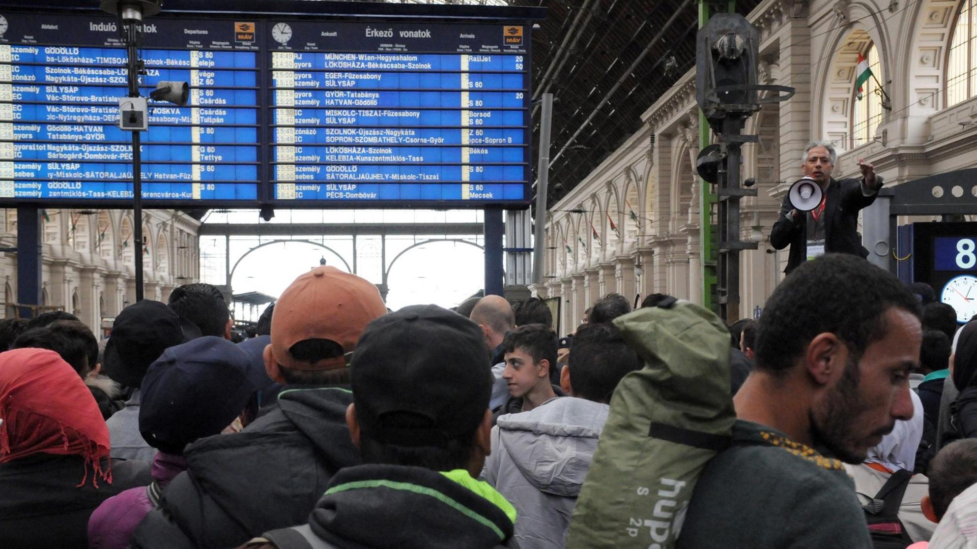 Flüchtlinge stehen auf dem Ostbahnhof in Budapest und hören einem Man mit Megafon zu.