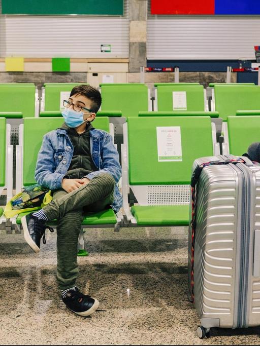 Ein junge sitzt mit Maske in einem Flugzeug-Terminal mit großen Gepäck (Symbolfoto)