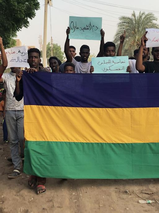 Jugendliche mit einer großen sudanesischen Fahne auf einer Landstraße halten Protestplakate hoch