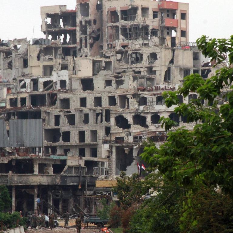 Ein durch Bomben zerstörter Wohnblock in Homs, Syrien im Mai 2014. Soldaten stehen vor der Ruine. Bäume säumen die Straße