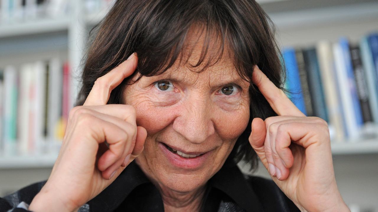 Die Kulturwissenschaftlerin Aleida Assmann, vor einem Bücherregal, hält sich ihre Zeigefingern an die Schläfe.