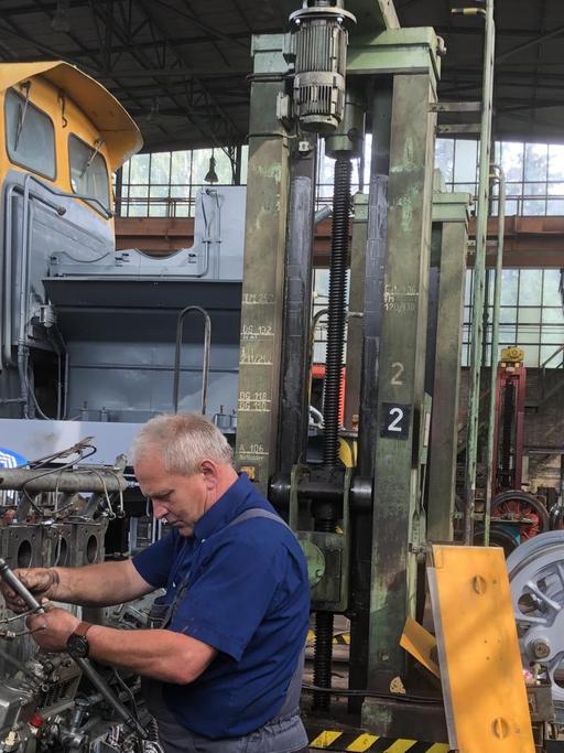 In einer Fabrikhalle steht ein Arbeiter an einer Lokomotive mit einem orangen Fahrerhaus