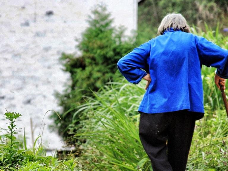Das Foto zeigt eine alte Frau in blauer Bluse mit einem Gehstock. Sie ist von hinten zusehen und steht in einem Garten.