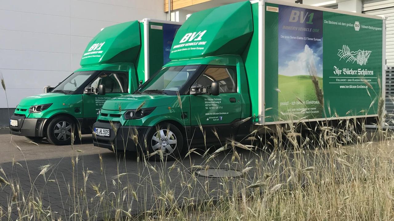 Zwei elektrisch angetriebene Lieferwagen der Bäckerei Schüren stehen auf dem Firmengelände. Sie sind in einem kräftigen Grün gestrichen, die Modellbezeichnung BV1 ist an mehreren Stellen deutlich zu lesen.