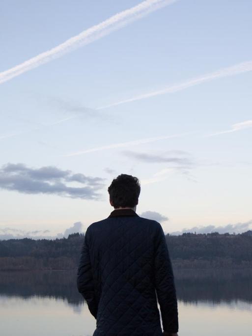 Ein Mann steht mit dem Rücken zum Betrachter vor einem See. Am Himmel sind Kondenstreifen zu sehen.