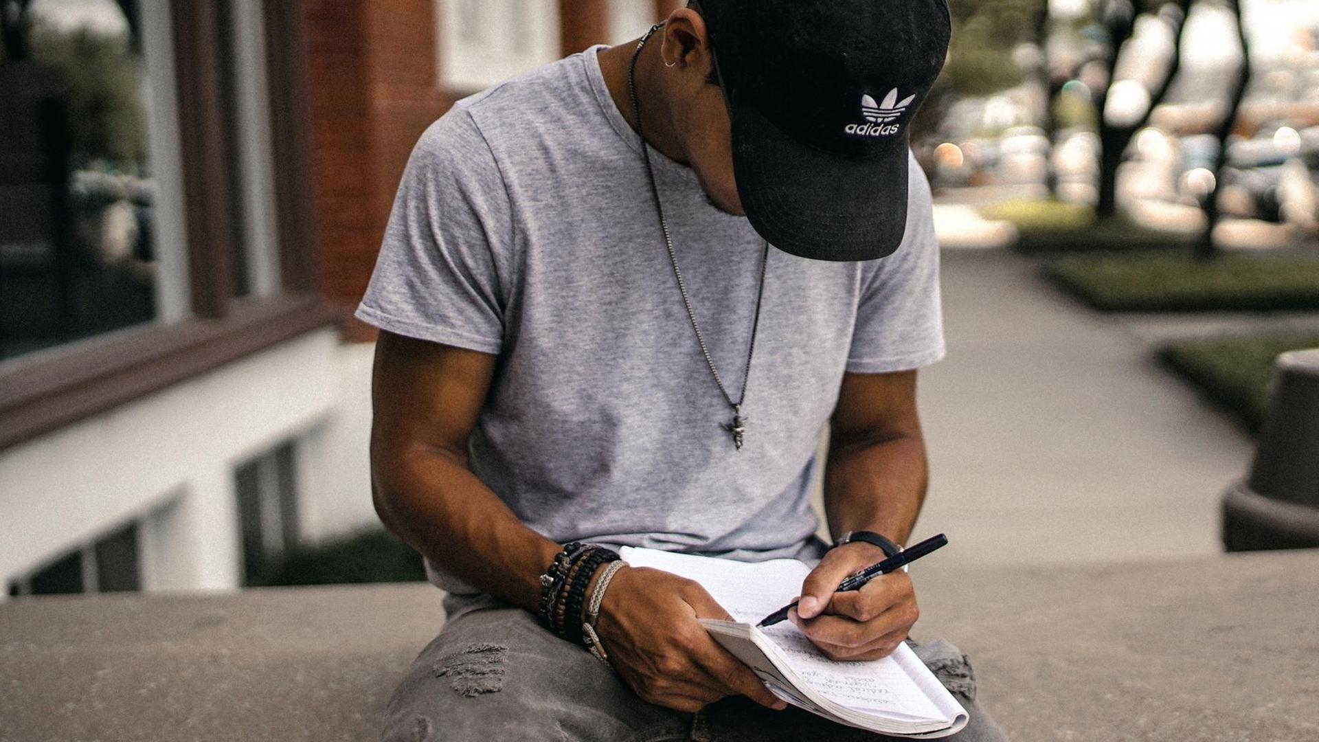 Ein junger Mann mit einer Basecap sitzt alleine vor dem Eingang eines Hauses und schreibt etwas in ein Heft.