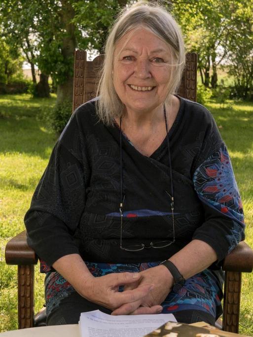 Helga Schubert sitzt auf einer Wiese in einem Stuhl, im Hintergrund sind Bäume zu sehen.