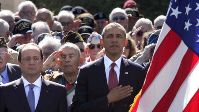 Hollande und Obama stehen nebeneinander, hinter ihnen eine Menschenmenge, Obama hält die Hand auf der Brust, im rechten Vordergrund verdeckt eine US-Flagge ein Drittel des Bildes