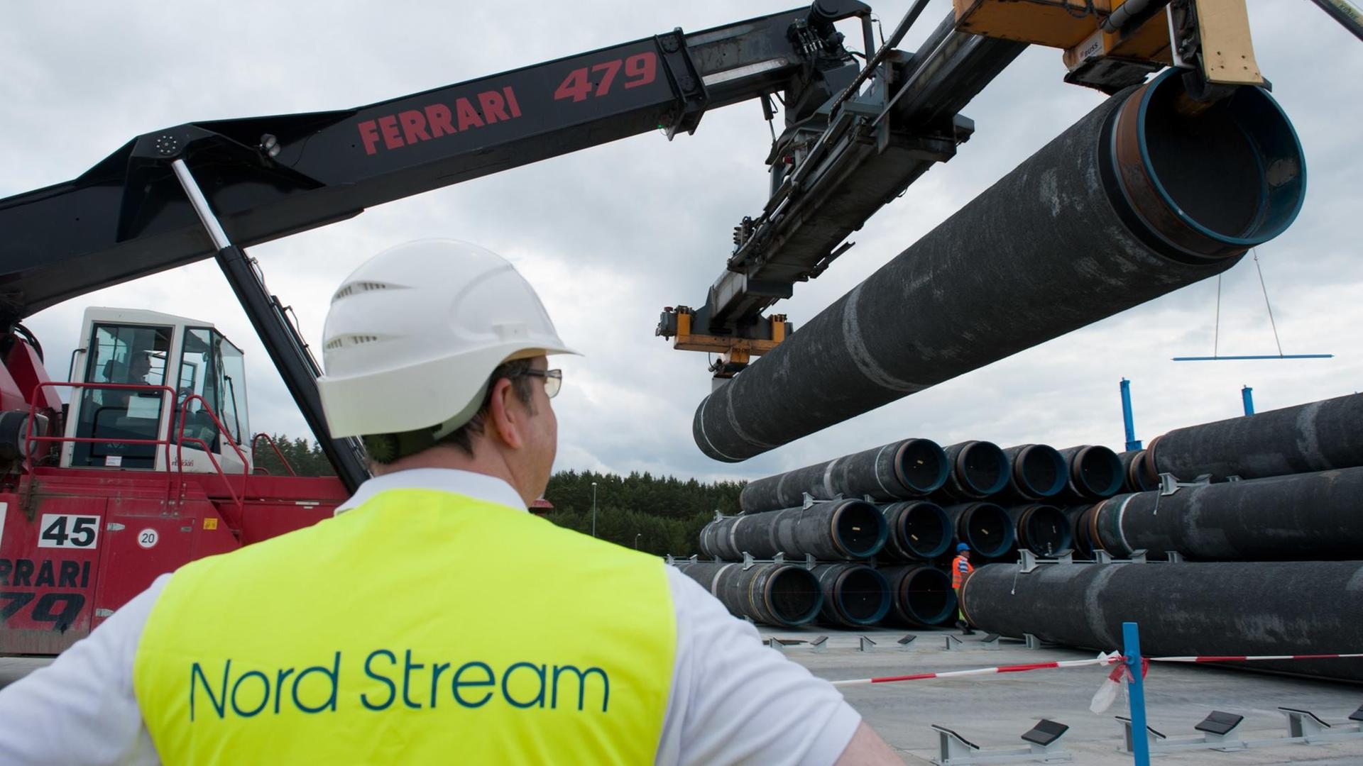 Im Vordergrund steht ein Mann in gelber Warnweste und beobachtet einen Bagger, der Pipelinerohre hebt.