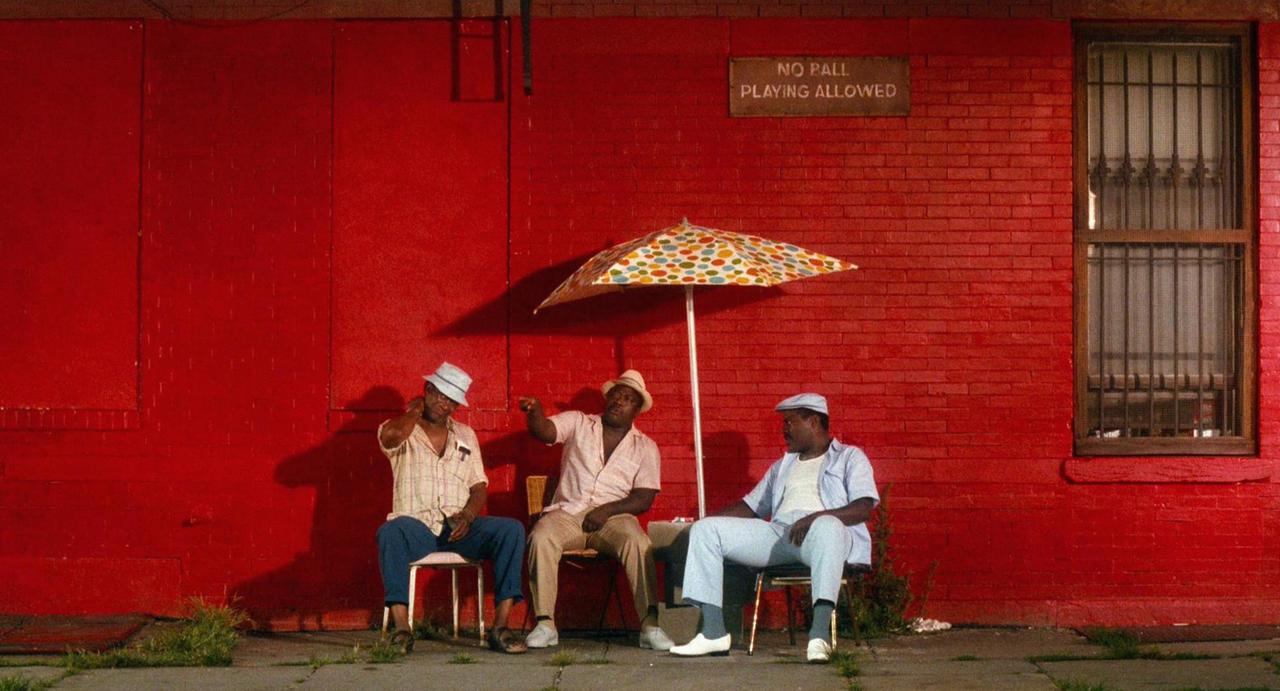 Szene aus dem Film "Do The Right Thing" von Regisseur Spike Lee aus dem Jahr 1989. Vor einer roten Hauswand sitzen bei glühender Hitze drei Männer unter einem Sonnenschirm.