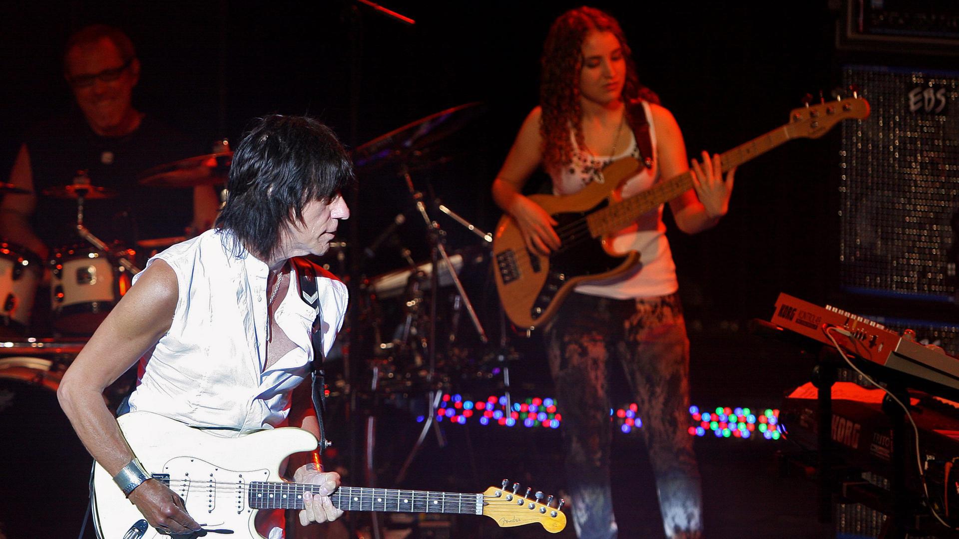 Ein weiß gekleideter Mann spielt auf einer weißen Gitarre, im dunklen Bühnenhintergrund steht eine Frau mit Baßgitarre.