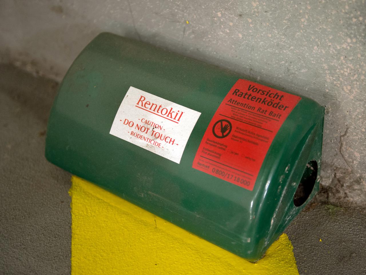 Eine grüne Box mit einem Loch an der Seite steht auf einem Asphaltboden, oben ein Aufkleber mit der Aufschrift "Vorsicht Rattenköder"