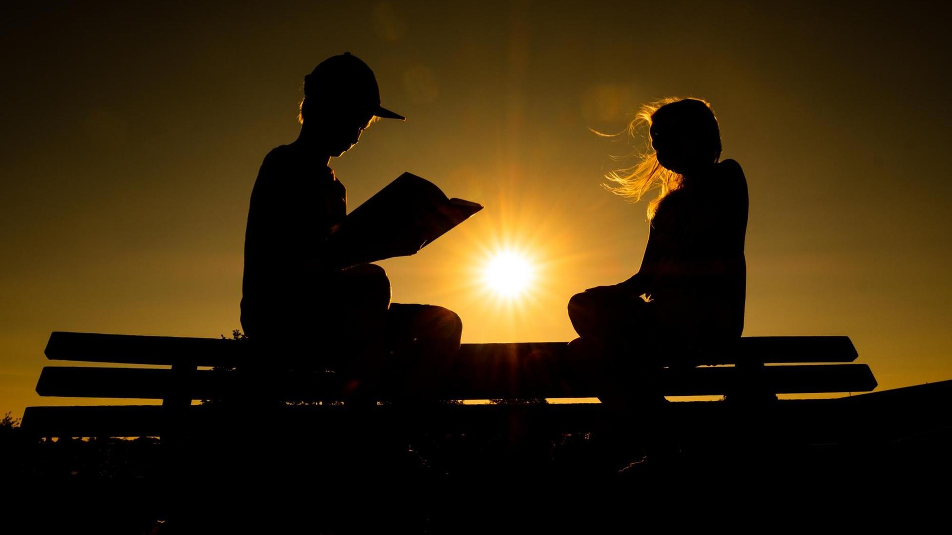 03.08.2018, Niedersachsen, Leer: Zwei Geschwister sitzen auf einer Parkbank und lesen, während hinter ihnen die Sonne untergeht. Foto: Mohssen Assanimoghaddam/dpa