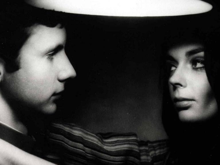 Filmszene, schwarz-weiß: Ein junger Mann und eine junge Frau stehen sich nah gegenüber und schauen sich an, ihre Arme um seinen Hals.