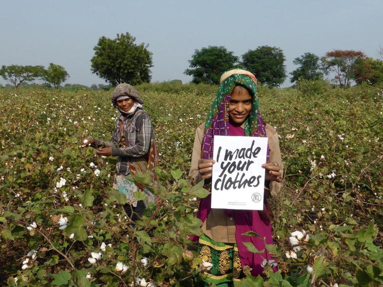Zwei Arbeiterinnen auf einem Baumwollfeld halten ein Schild hoch auf dem steht "ich habe deine Kleidung gemacht".