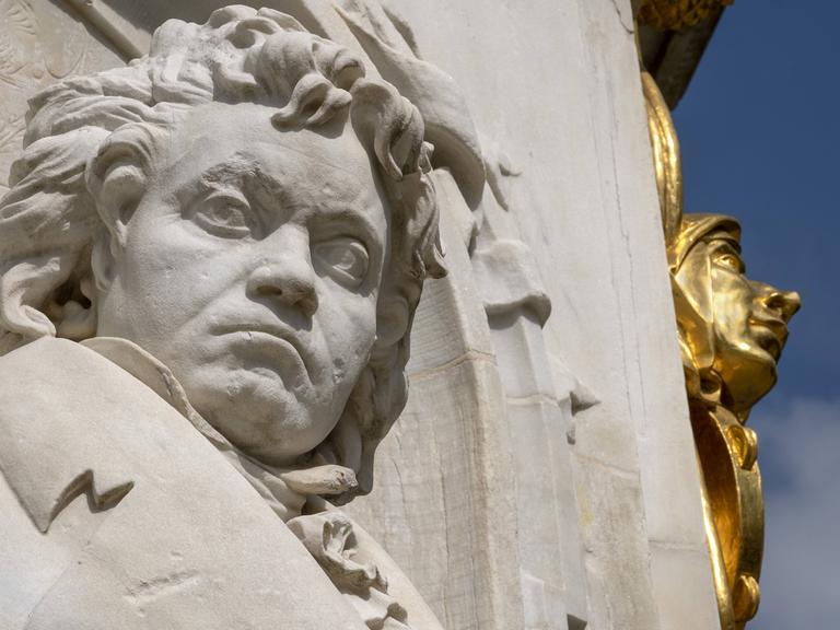 Ein Denkmal von Ludwig van Beethoven aus hellem Stein, zu sehen ist er von Kopf bis Brust, im Hintergrund ein vergoldeter Teil des Denkmals und blauer Himmel.