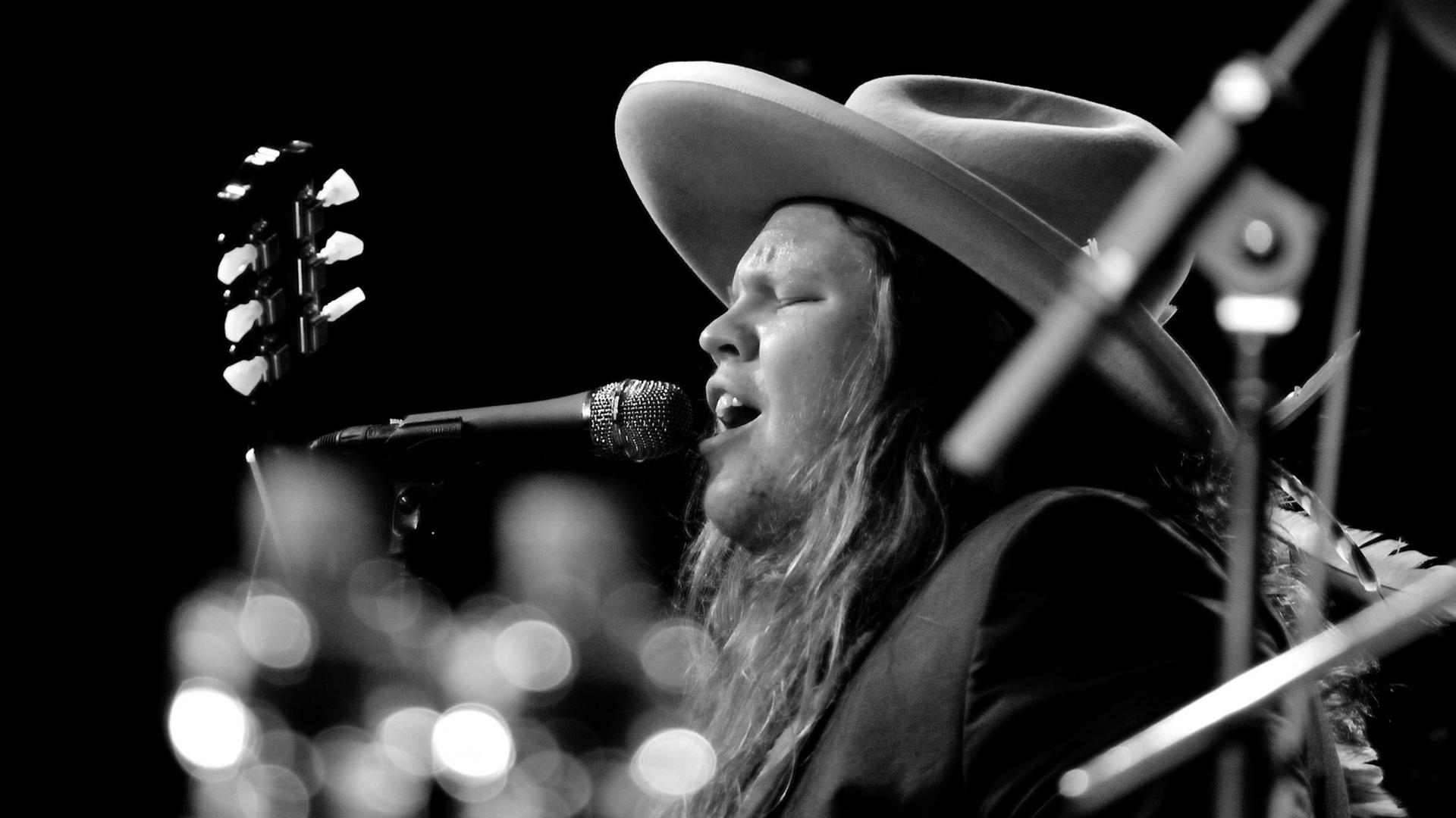 Ein junger Mann mit blonden langen Haaren steht auf einer Bühne, spielt Gitarre und singt, dabei trägt er einen großen runden Hut.
