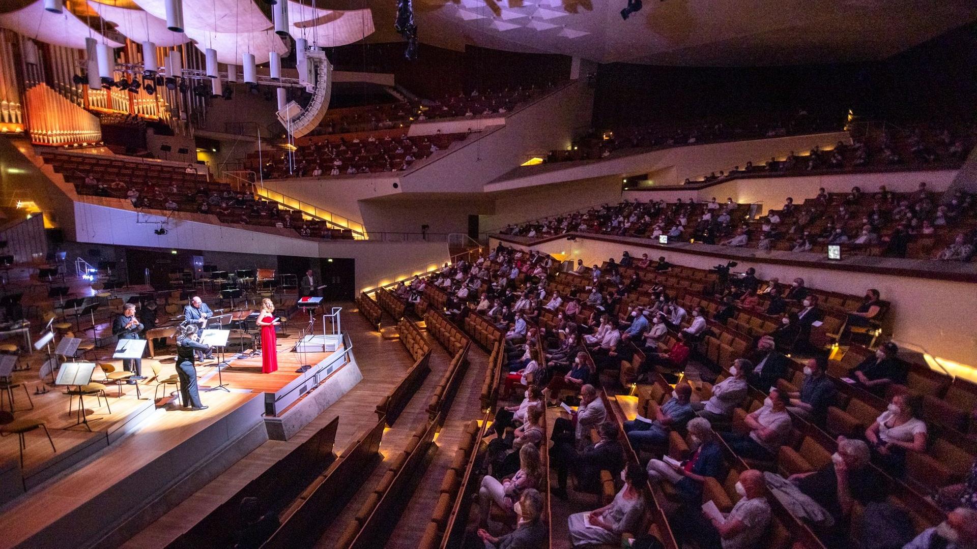 In einem großen Konzertsaal mit reduzierter Publikumsbesetzung spielen links auf der Bühne drei Musiker hinter einer Sängerin im roten Kleid.