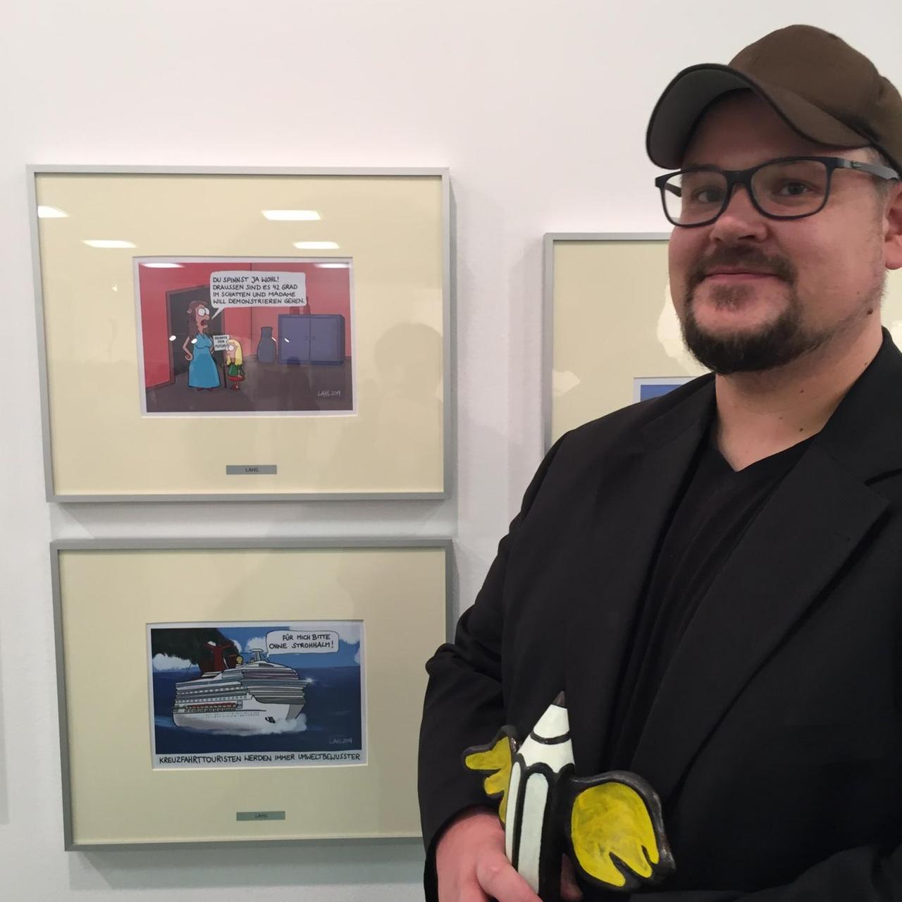 Der Newcomer-Gewinner des Deutschen Karikaturenpreises: Lars Murach alias Lahs steht mit dem Geflügelten Bleistift vor seinen Cartoons