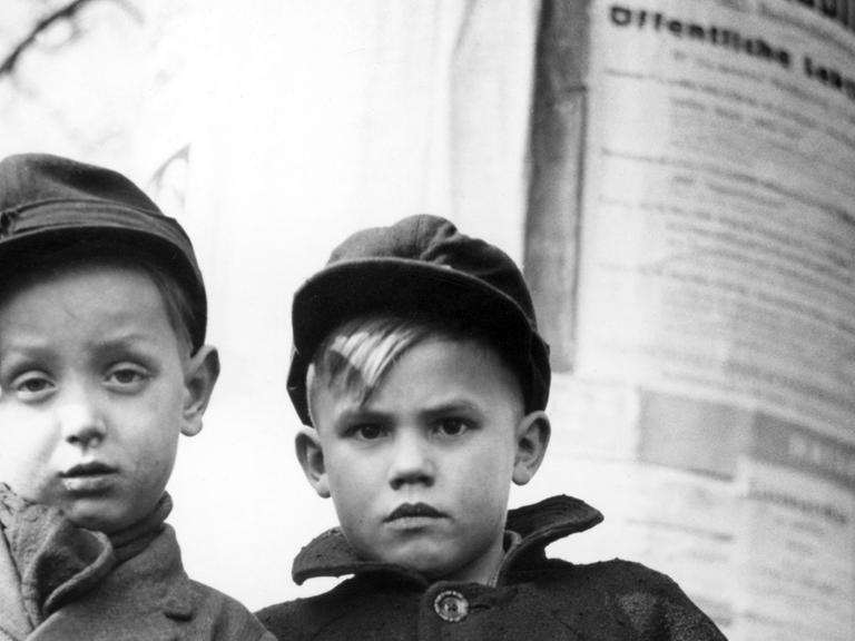 Zwei Berliner Jungs stehen mit skeptischem Gesichtsausdruck und den Händen in den Taschen vor einer Lifaßsäule. Aufnahme aus den 50er-Jahren.
