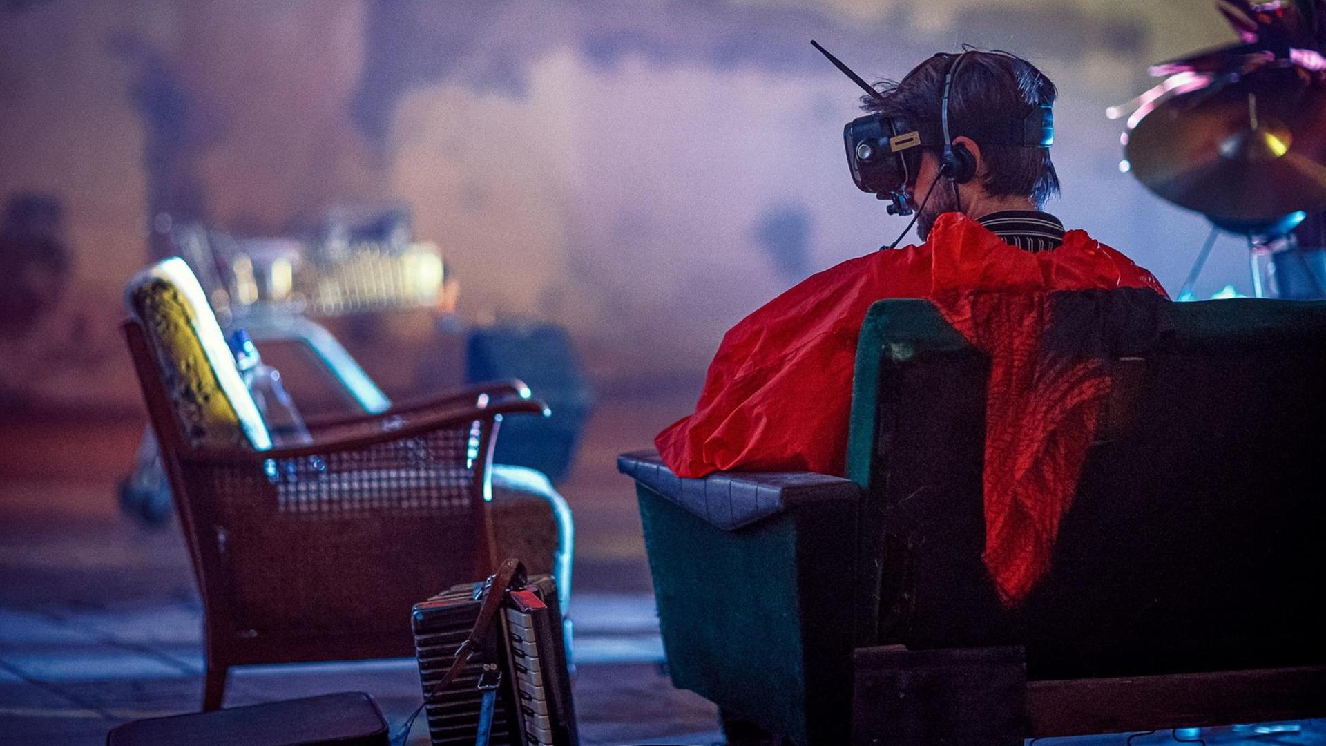 Ein Mann ist von hinten zu sehen, wie er auf einen Sessel sitzt, eine VR-Brille samt Antenne auf dem Kopf trägt und ein auffallend rotes Oberteil. Neben ihm auf dem Boden steht ein Akkordeon. Ein Mann ist von hinten zu sehen, wie er auf einen Sessel sitzt, eine VR-Brille samt Antenne auf dem Kopf trägt und ein auffallend rotes Oberteil. Neben ihm auf dem Boden steht ein Akkordeon.