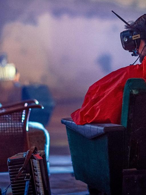 Ein Mann ist von hinten zu sehen, wie er auf einen Sessel sitzt, eine VR-Brille samt Antenne auf dem Kopf trägt und ein auffallend rotes Oberteil. Neben ihm auf dem Boden steht ein Akkordeon.