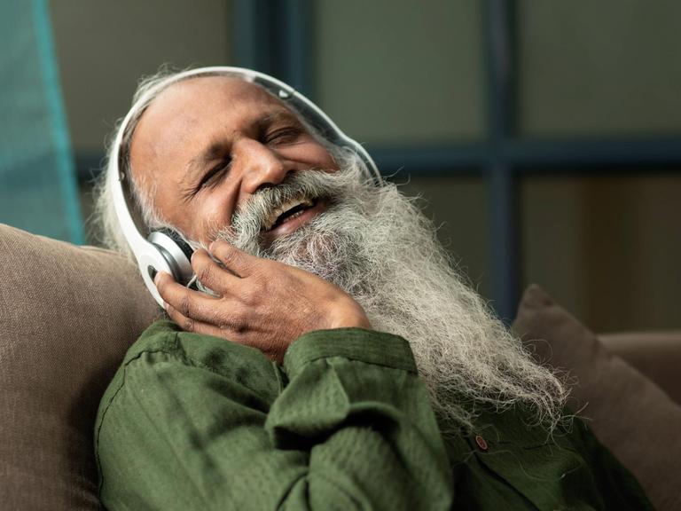 Ein Mann mit weißem Bart hört lachend und glücklich etwas über Kopfhörer.