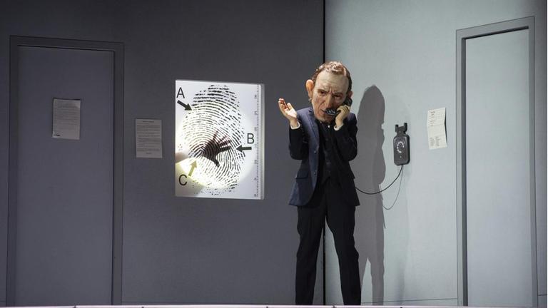 Ein Mann mit einer überdimensionierten Kopf-Maske telefoniert in einem grauen Zimmer. Hinter einem Screen, auf dem ein Fingerabdruck zu sehen ist, sieht man eine Hand.