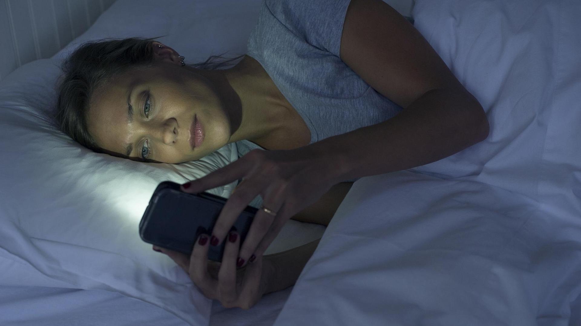 Eine Frau liegt im Dunkeln im Bett und schaut auf das leuchtende Display ihres Smartphones