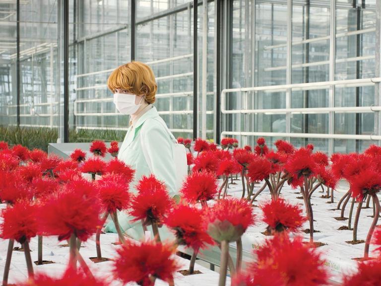 Die Genetikerin Alice (Emily Beecham) mit Mundschutz im Labor, umgeben von roten Blumen.