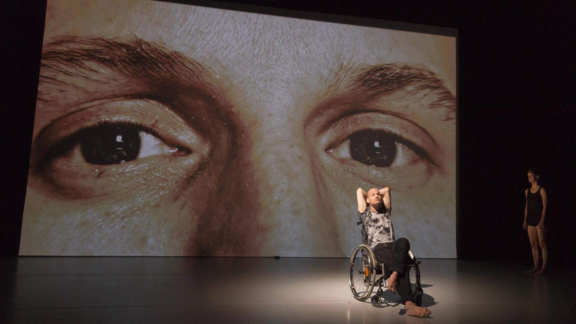 Ein Mann sitzt auf der Bühne in einem Roll-Stuhl. Hinter ihm hängt ein riesiges Bild mit 2 Augen. 