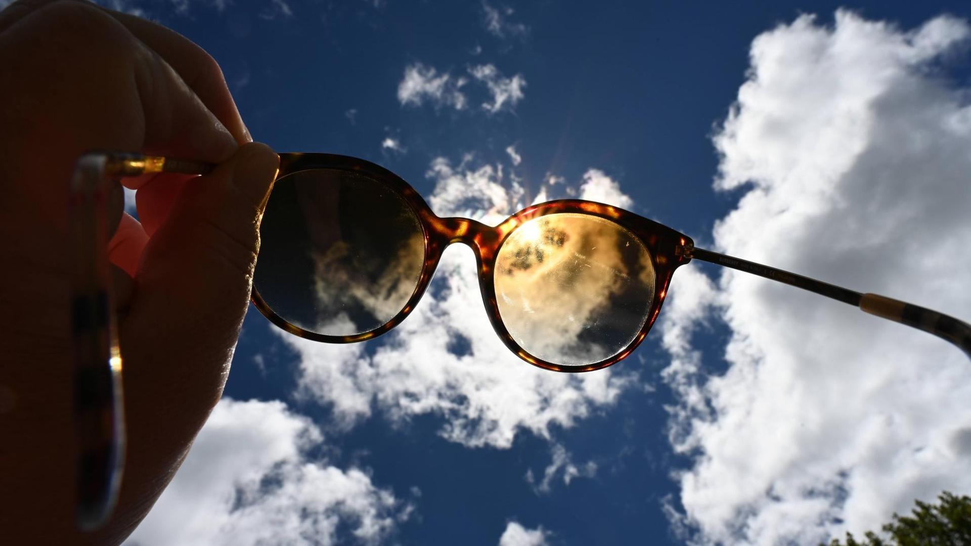 Eine Sonnenbrille vor dem blauen Wolkenhimmel mit der Sonne.
