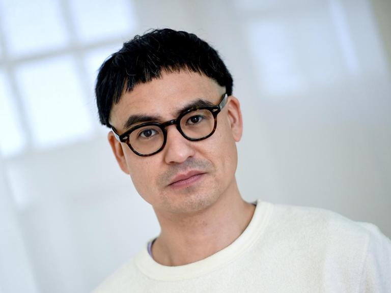 Portrait des Regisseurs Burhan Qurbani, im weißen Pullover vor weißen Vorhängen.
