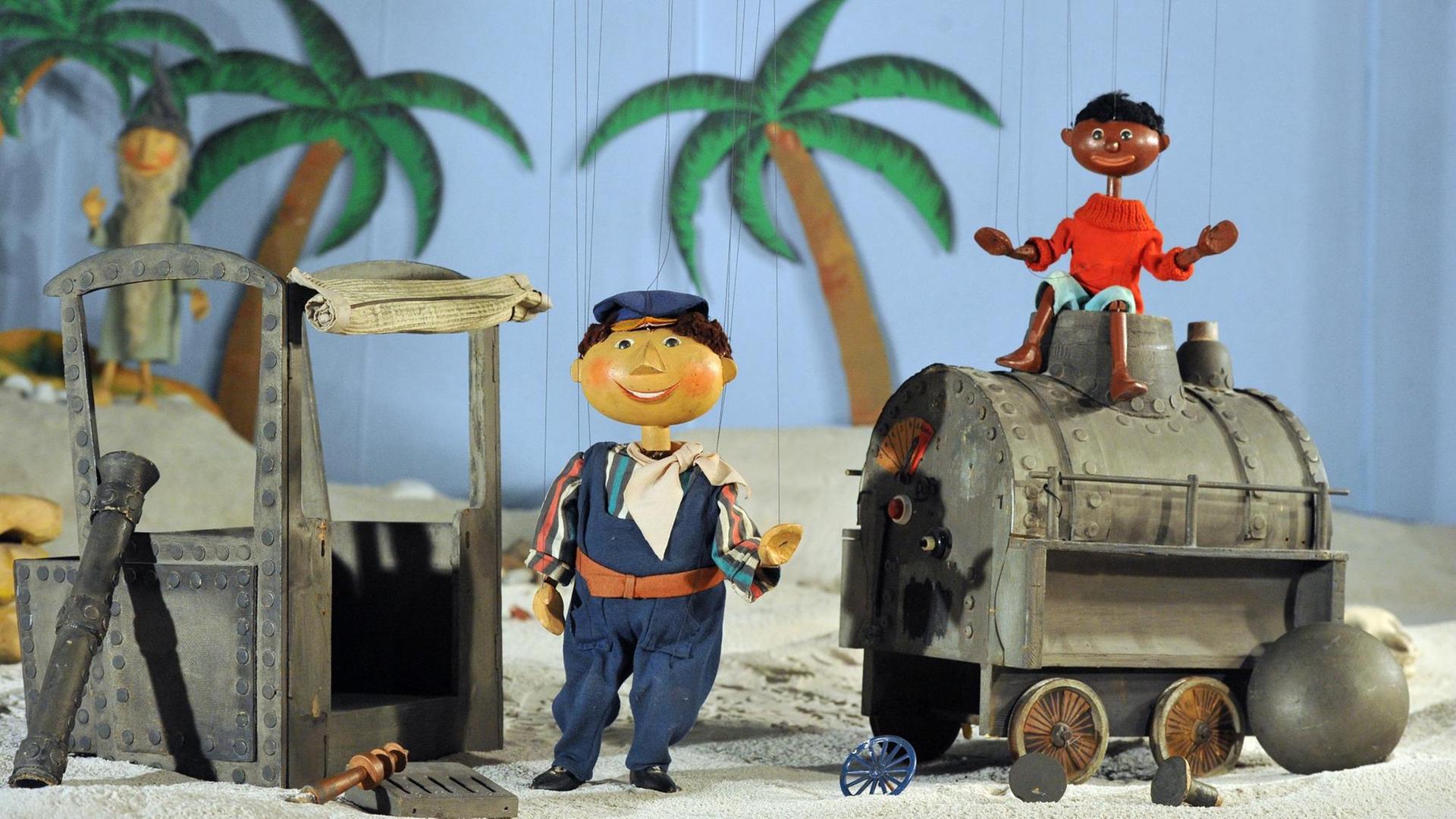 Die Marionetten Scheinriese Tutur, Lukas und Jim Knopf stehen in Augsburg im Puppenkistenmuseum mit der zerstörten Lokomotive Emma in einem Wüstenszenario.