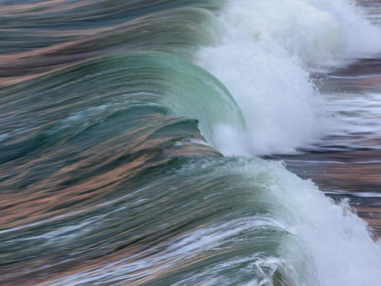 Seitlicher Blick auf ein lange Welle aus blau-grünem Meereswasser mit rötlichen Reflexionen auf der Oberfläche, die sich gerade überschlägt und weiße Gischt bildet.