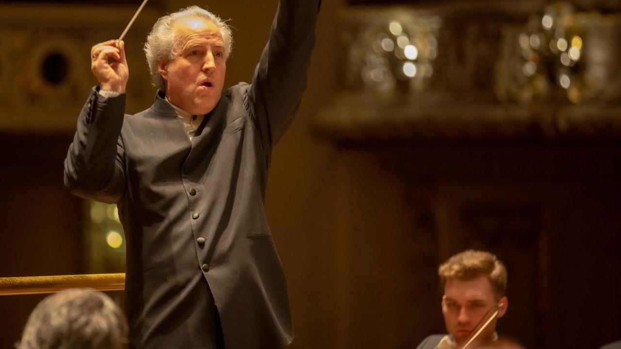Ein älterer Herr mit weißem, nach hinten gekämmtem Haar streckt Taktstock und Arme nach oben, um ein Orchester zu dirigieren.