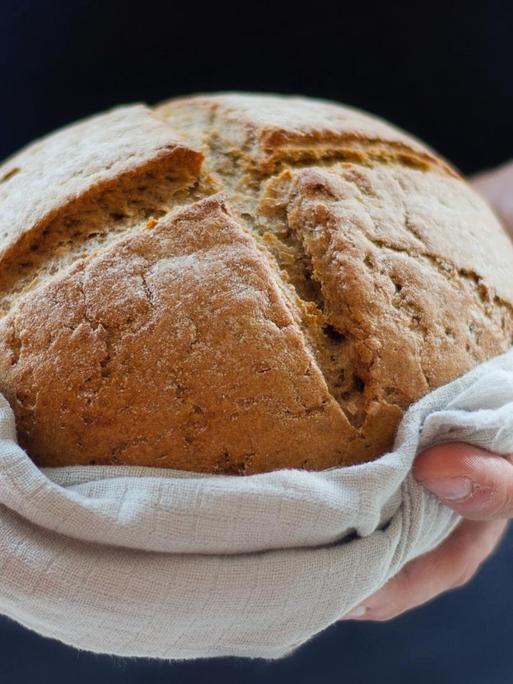 Ein frisch gebackenes Brot in einem Leinentuch von zwei Händen gehalten.