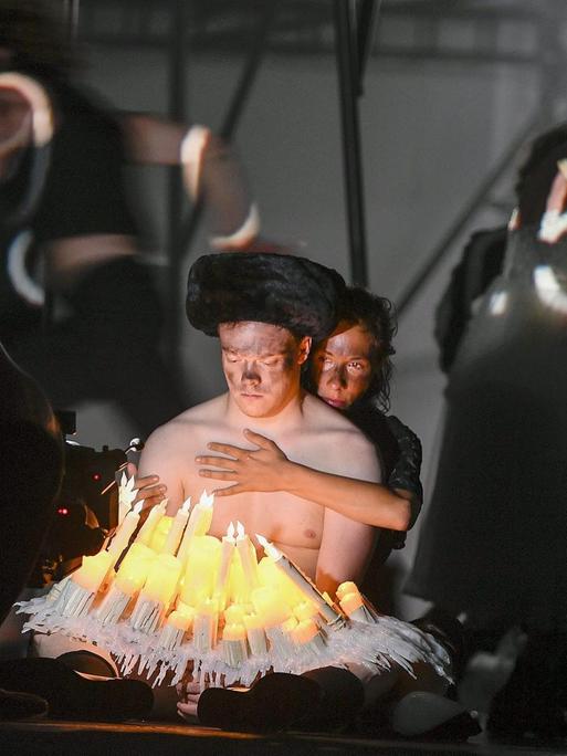 Ein Mann mit nacktem Oberkörper sitzt vor mehreren elektrischen Kerzen. Eine Frau sitzt hinter ihm und umarmt ihn.