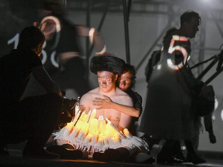 Ein Mann mit nacktem Oberkörper sitzt vor mehreren elektrischen Kerzen. Eine Frau sitzt hinter ihm und umarmt ihn.