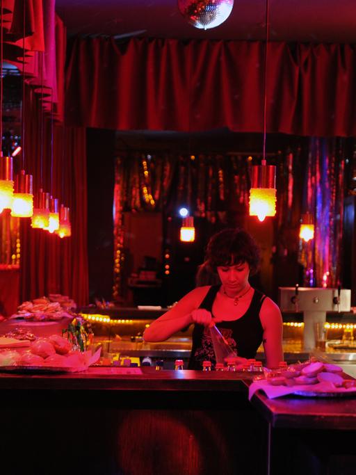 Eine Bar in Berlin, in rotes Licht getaucht. Eine Frau hinterm Tresen, zwei Menschen an der Bar.